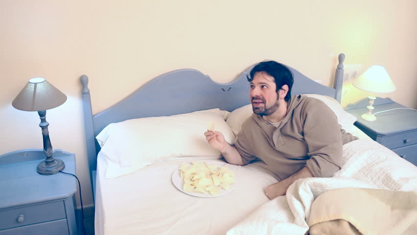 A man lies in the bed, he's recovering from a bad flu / cold / illness. A woman (his wife) is near him. The couple eats tasty potato chips.
