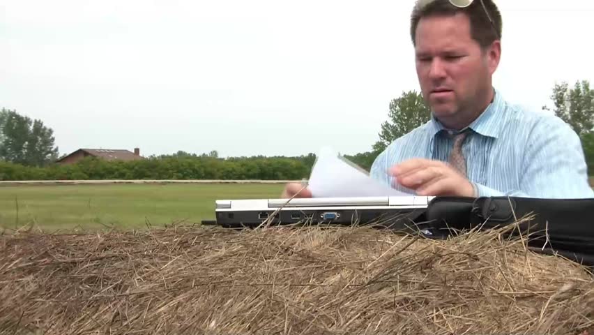 Businessman on hay bail in rural setting writes with marker on laptop until he's done, and turns white paper around reading need more customers.