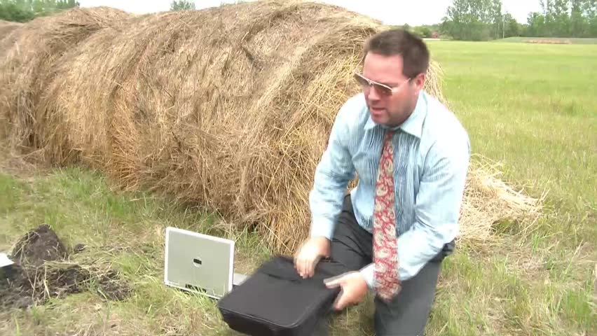 Businessman's foot is stuck in hole in rural setting as he pleads for help while still trying to get leg out, crooked sunglasses, hay bail and laptop computer in frame.