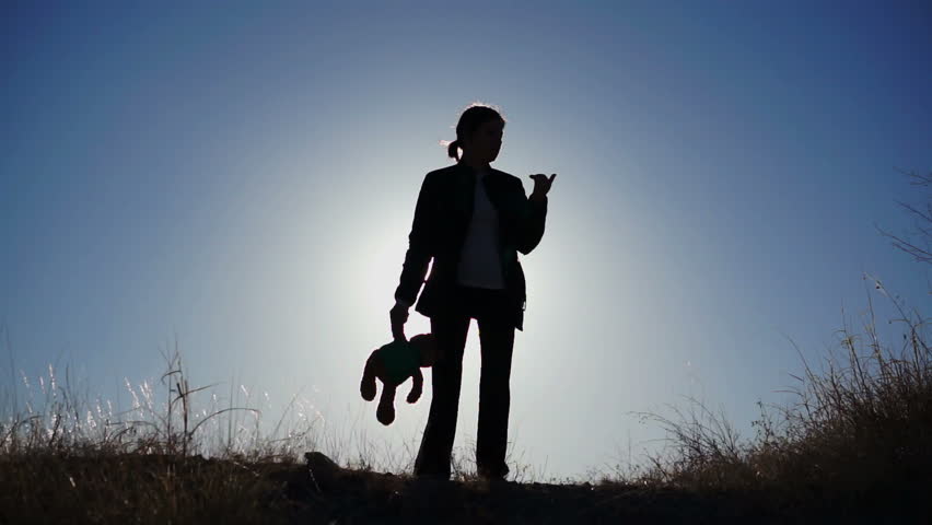 Silhouette of an innocent looking, lonely girl holding her teddy bear in one hand against a clear blue sky on a breezy day.
