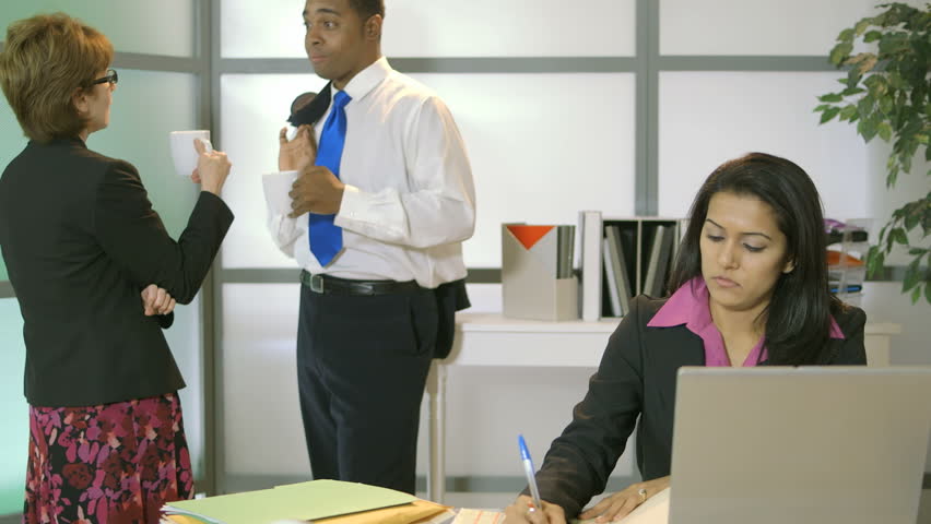 A lovely woman working at her computer is annoyed by the office gossip going on behind her. 