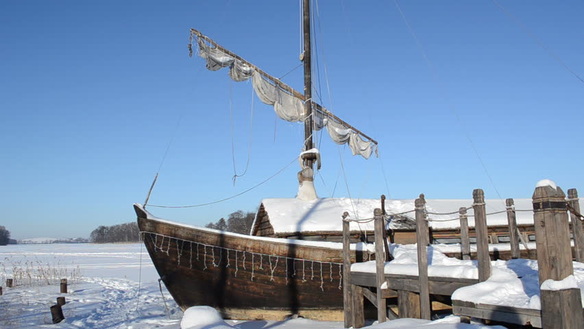 retro wooden ship frozen in lake ice near pier and sail move in wind on background of blue sky.