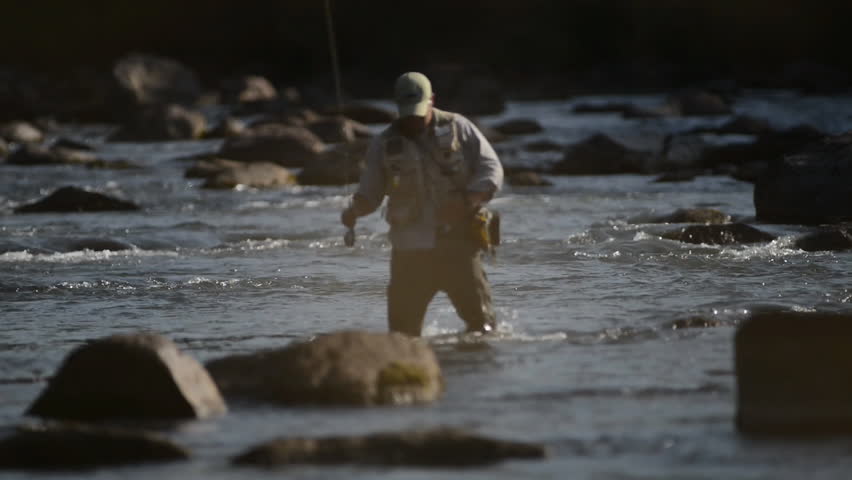 A man in waterproof overalls stands in a river and casts his lure with his rod while he spends his day fly fishing.