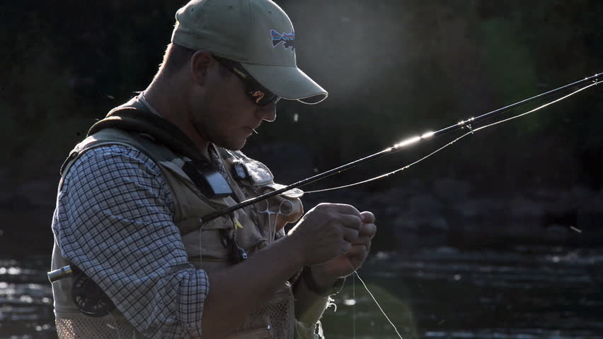 A man in waterproof overalls stands in a river and casts his lure with his rod while he spends his day fly fishing.