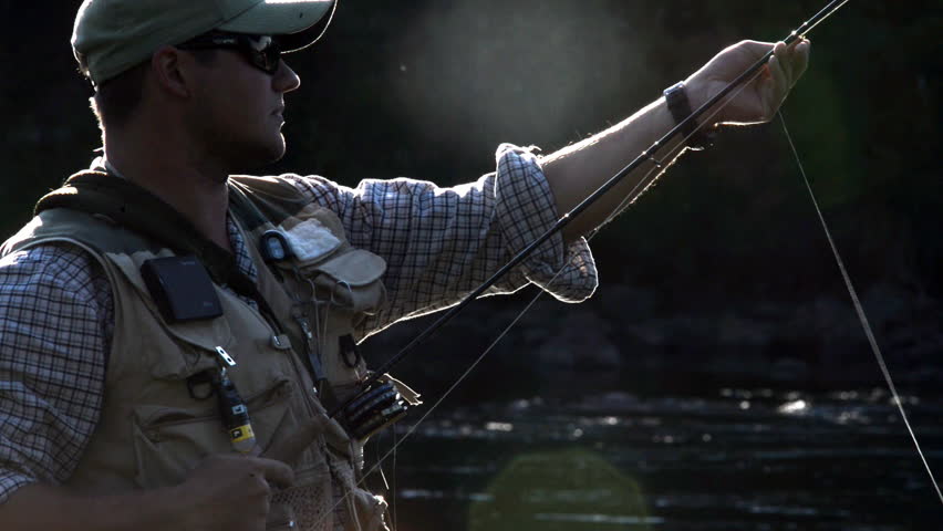 A man in waterproof overalls stands in a river and casts his lure with his rod while he spends his day fly fishing.