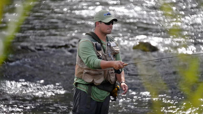 A man in waterproof overalls stands in a river and casts his lure with his rod while he spends his day fly fishing.