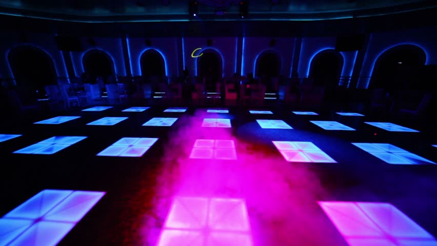 People sit at tables near dance floor with color light and smoke in night club