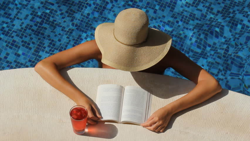 Woman with wide brimmed hat reads by the side of a pool. Tulum, Mexico.