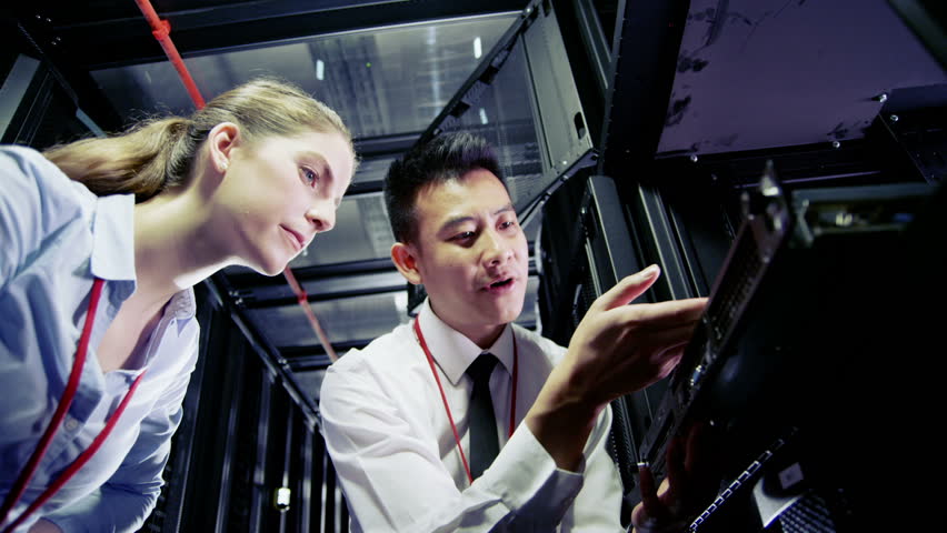 Two IT engineers are working in a data center with rows of server racks and super computers. They are discussing their work as they check cables and other equipment. In slow motion.
