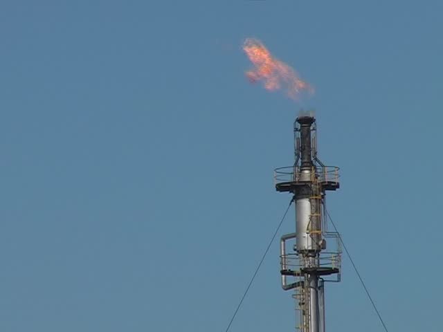 Gas flares and burn off from a chimney at a fuel plant on a sunny day against a blue sky, 