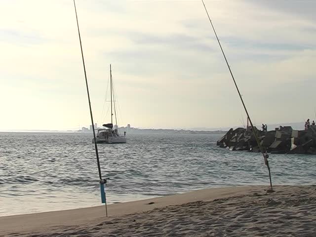 Catamaran sailing away on the ocean, after sunset into the harbor. Fishing rods on the beach in the foreground