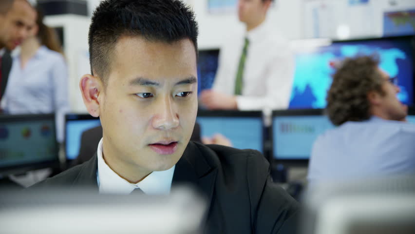 Young and ambitious stock market trader is doing a deal over the phone in a busy office filled with computers. The rest of his team are hard at work in the background. 