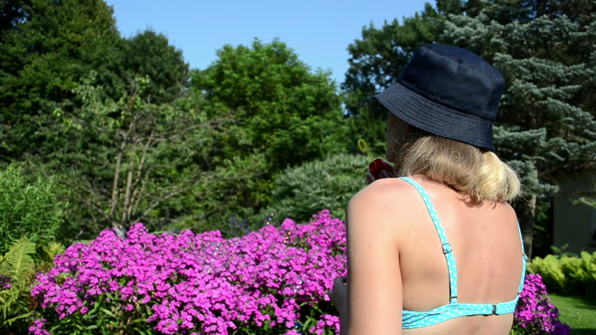 young woman with bra and dark cap blow soap bubbles on background of red flowers in garden.