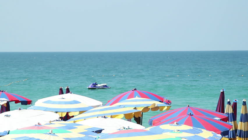 Sunny beach umbrellas - Summer video background featuring vivid striped multicolor beach umbrellas in focus and a jetski on waves in the distance