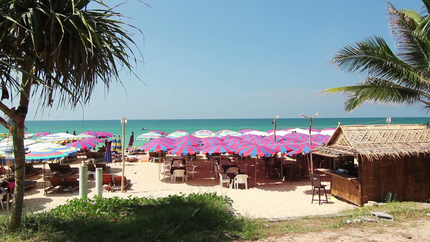 Hot tropical Summer day - Idyllic tourist resort scene with colorful beach umbrellas, sunbeds, and a small palmtree beach bar for drinks at the beach. Horizon in far distance, blue sky, gentle breeze