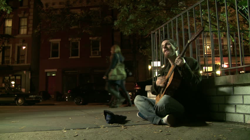 Young man playing a worn-out guitar on the night time streets of Manhattan, NY. Young woman approaches, listens for a while then leaves some money. 