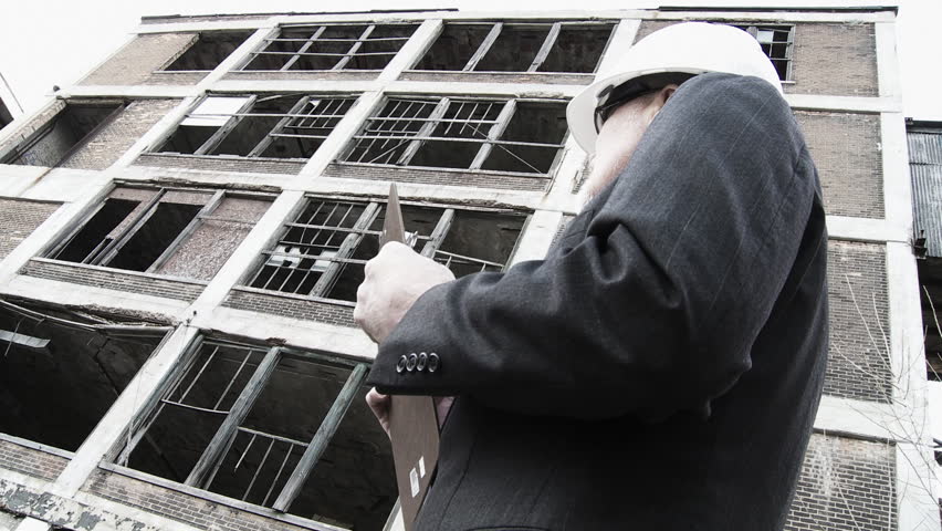Developer walks into shot and looks around at the collapsed buildings around him, making notes. Low, wide angle view with high contrast and low color-saturation.
