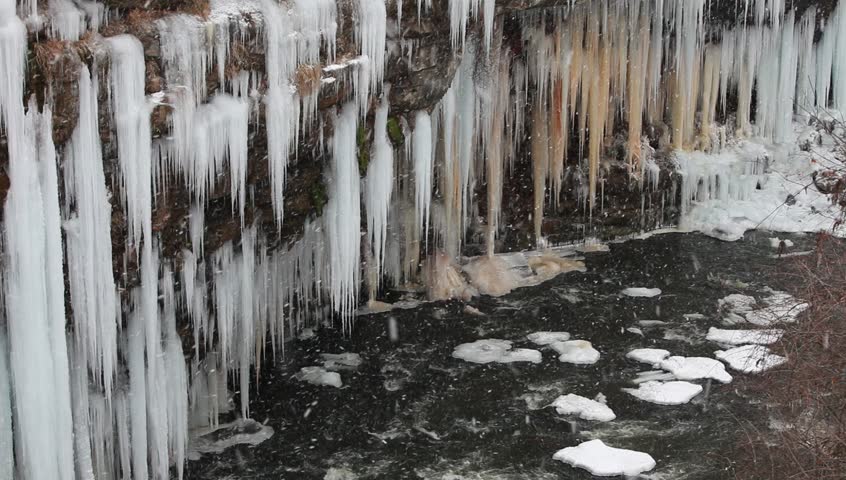 icicles on a cliff wall hanging over a flowing river with snow flakes falling.