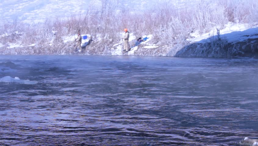 Two fisher men can bee seen standing on a snowy river bank, at a distance.