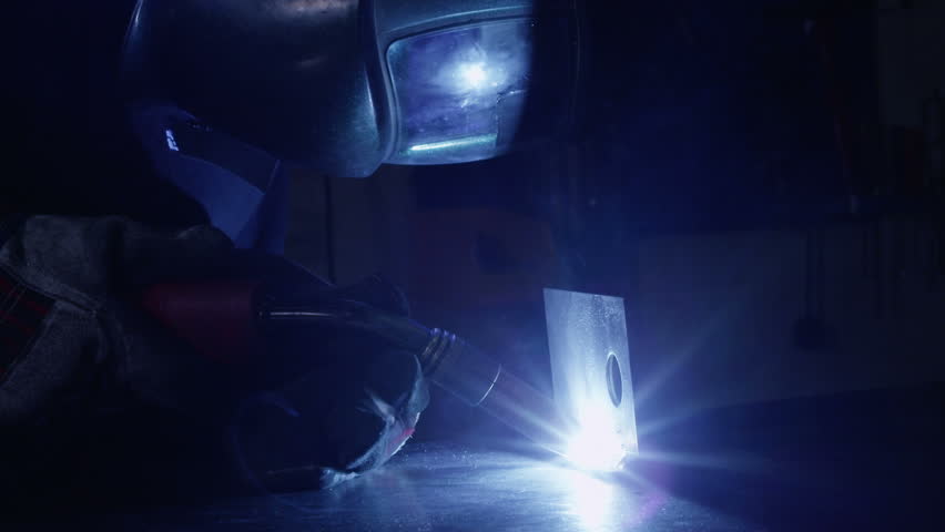 A welder is working in a dark construction or engineering plant. The flare of the flame is reflected in the visor of his mask.