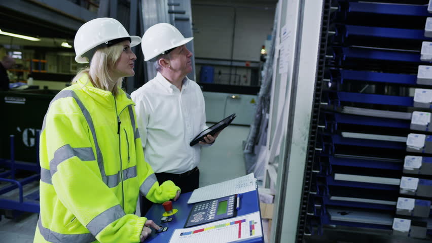A female worker within a factory or warehouse is being shown by her foreman how to operate some heavy machinery.