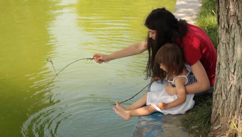 Brunette young adult mother teaches little baby daughter fishing.
