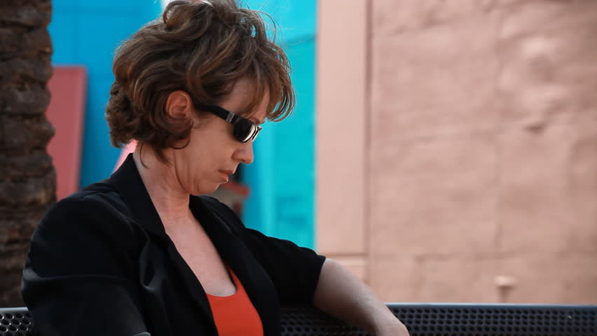 An annoyed or impatient woman sitting on a bench calling someone on her cell phone.