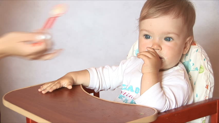 Baby sits at a children's little table. Mum feeds the child with porridge. Isolated on the white