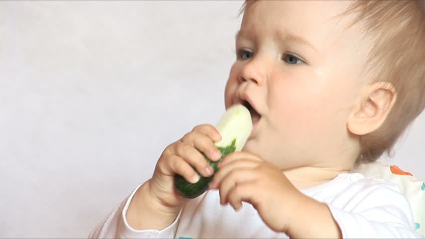 Baby sits at a table and eats a cucumber. Isolated on the white