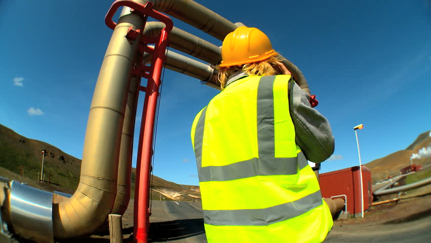 Female geothermal energy technician using a mobile (cell) phone while surveying the power plant