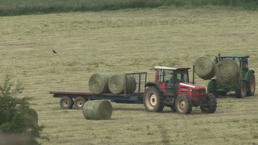 Tractor loads bales of hay onto a trailer as the baler passes. HD.