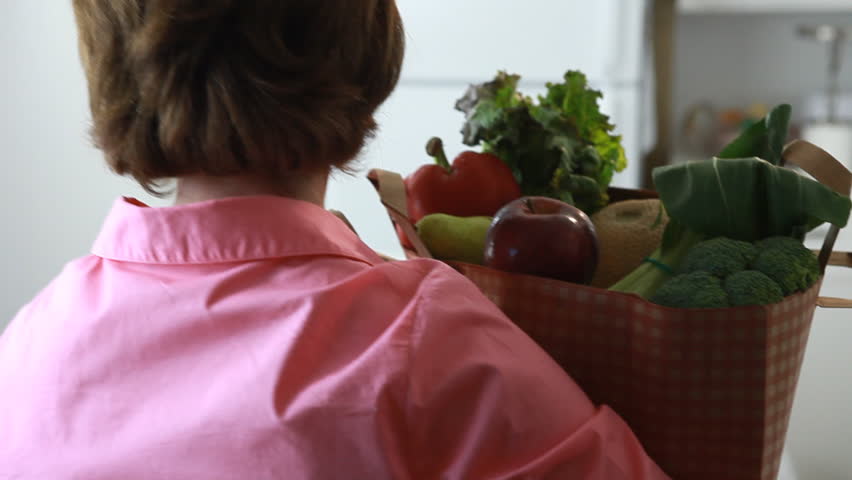 A woman carrying a grocery bag, places the bag on her kitchen counter and proceeds to take from the bag an assortment of fruits and vegetables. (Shoulder mount)