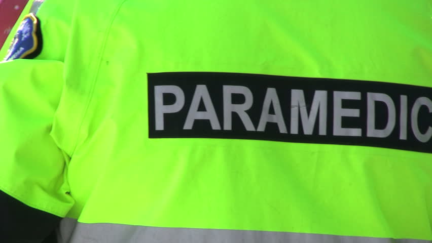 A paramedic wearing a black and green jacket stands by offering assistance during a crisis.