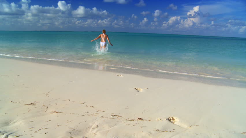 Young female in swimwear running across white sandy beach to swim in aquamarine waters 60 FPS