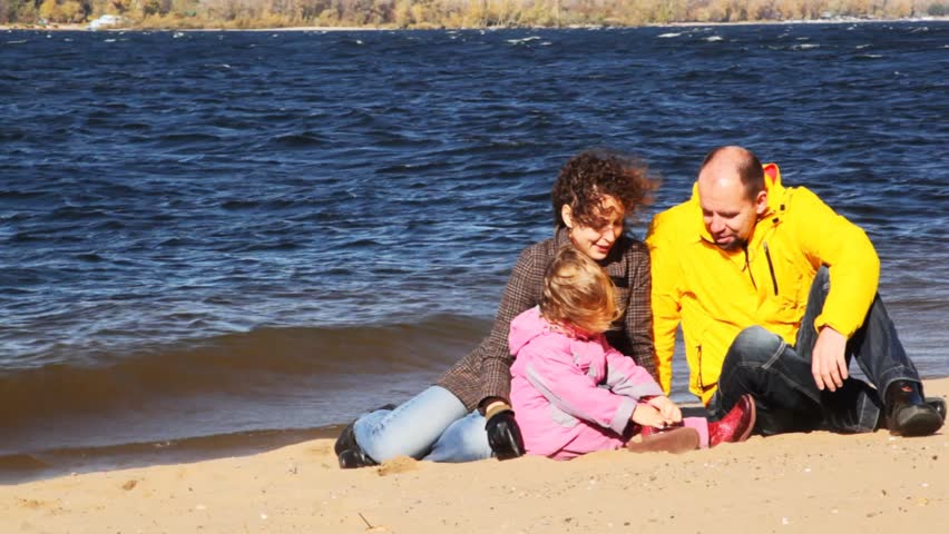 family of mum, daddies and daughter sits on sandy to river bank talk and smile 