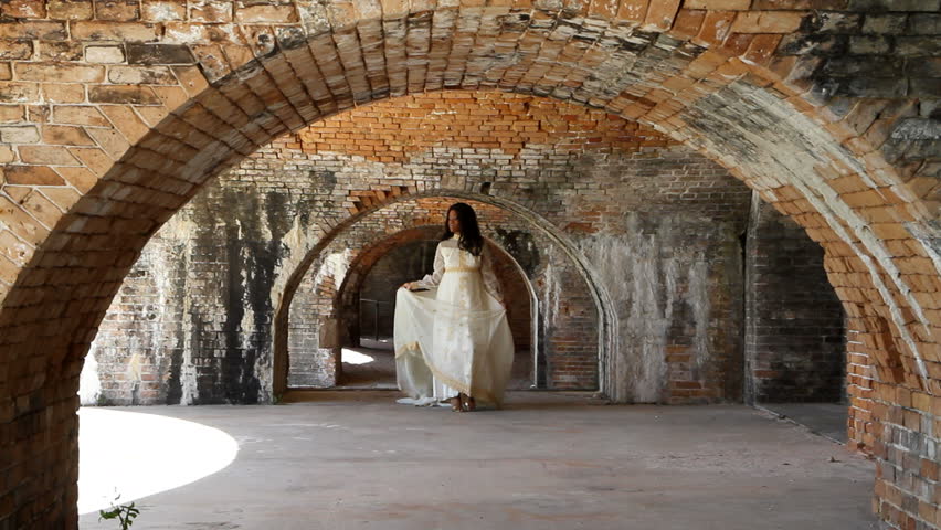 Black woman in a 1960's era bridal gown walks under the brick arches of an old fort built in the 1850's.