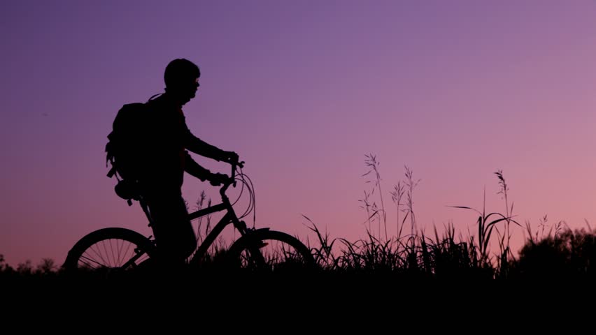 silhouettes of two bicyclists with knapsacks against sunset sky 
