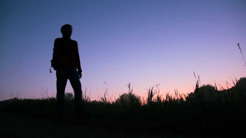 traveller with rucksack walks against sunset sky, stops, looks about and comes 