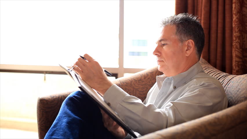 A relaxed man sitting by a big bright window in a large lounge chair finishes writing something in a notebook.