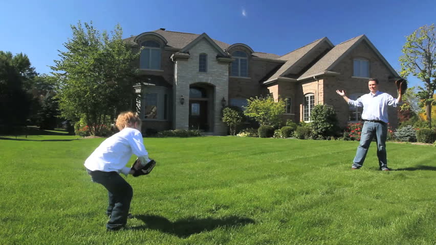 A father and young son play catch in front of their luxury home 