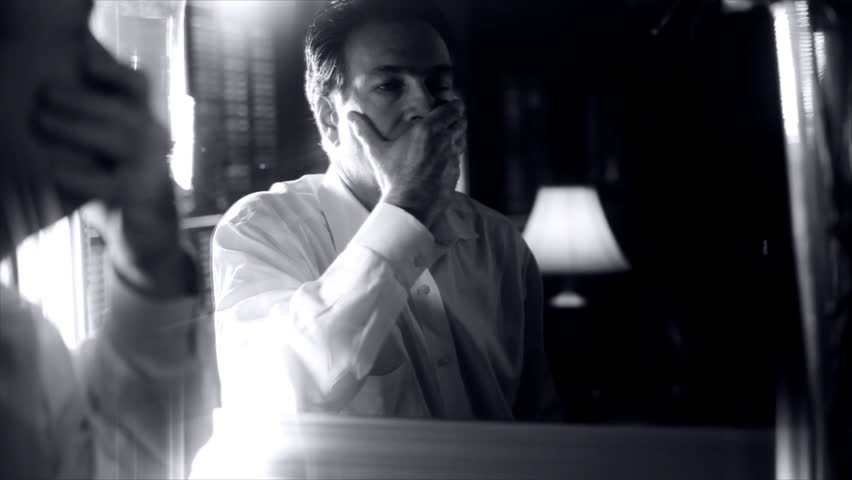 A distressed man standing in front of a mirror looks with fearful eyes at the person he sees looking back at him. Black and White with dreamy haze added for affect.