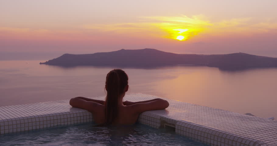 Woman In Swimming Pool Spa Enjoying Sunset On Over Sea Luxury Vacation Travel On Santorini, Greece, Europe. Girl in bikini watching beautiful view of sunset enjoying her holidays at resort hotel.