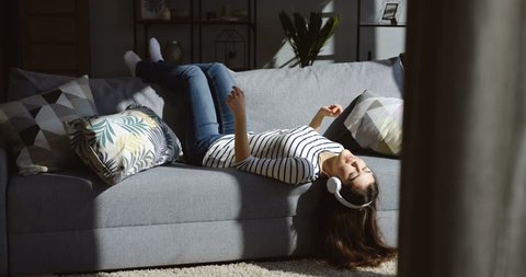 Young Cute Brunette Woman In The Big Headphones Lying On The Gray Sofa Upside Down Listening To The Music Singing And Moving In The Rhythm In The Cozy Living Room Indoors