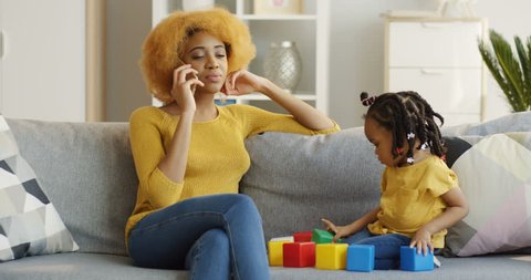 480px x 253px - Young african american mother talking on the phone while siting on the gray  sofa in the living room and her cute small daughter playing with colourful  cubes beside. indoors