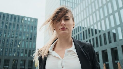 Portrait of young business woman in suit with blonde hair blowing in wind  walking on the urban business district street. skyscraper background.  business, finance, banking. girl looking at camera.