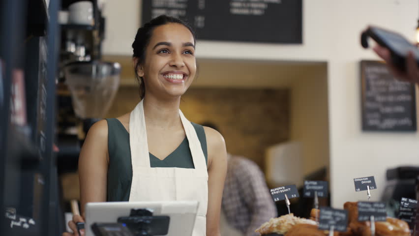 4K Cheerful worker serving a customer who uses smartphone to pay in coffee shop. UK - April, 2016